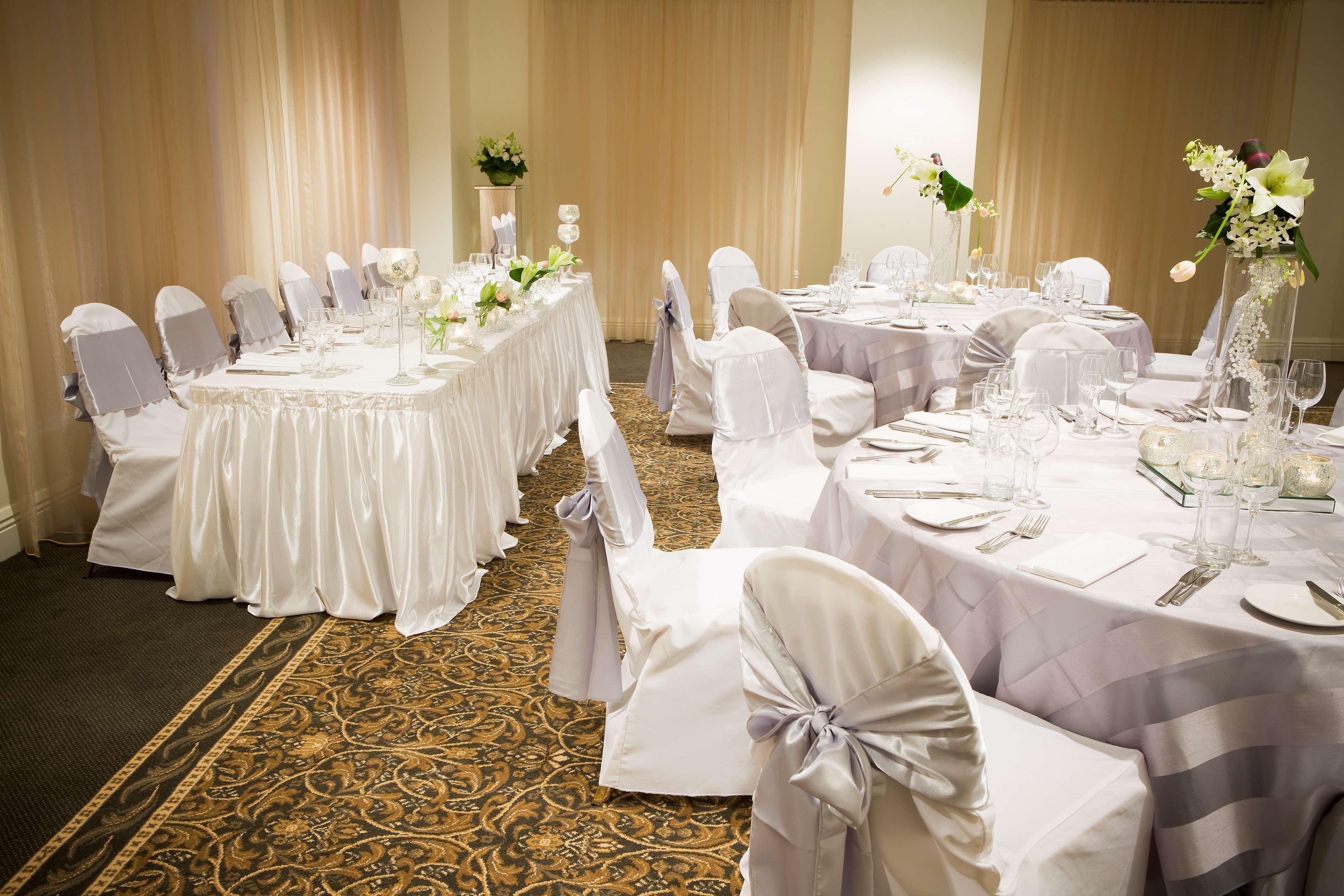 מלון Stamford Plaza מלבורן מראה חיצוני תמונה The photo depicts an elegantly set dining area, likely for a special event or celebration. There are several round tables covered with tablecloths, each set with plates, glassware, and cutlery. The chairs are dressed in white covers, some adorned wit