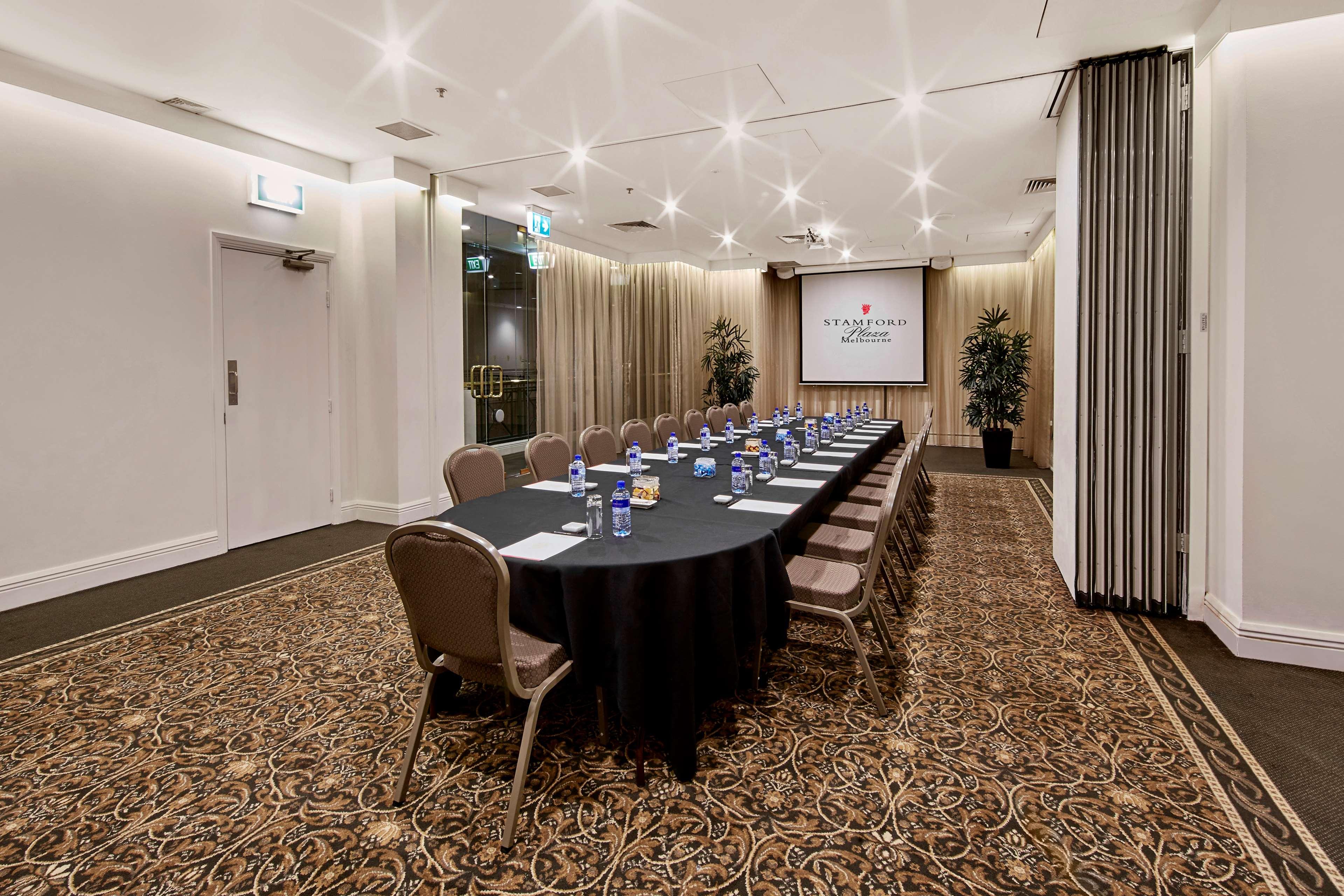 מלון Stamford Plaza מלבורן מראה חיצוני תמונה The photo shows a meeting room set up for a conference or presentation. There is a long, rectangular table covered with a black tablecloth, surrounded by chairs. Each seat has a water bottle placed in front of it. At the front of the room, there is a