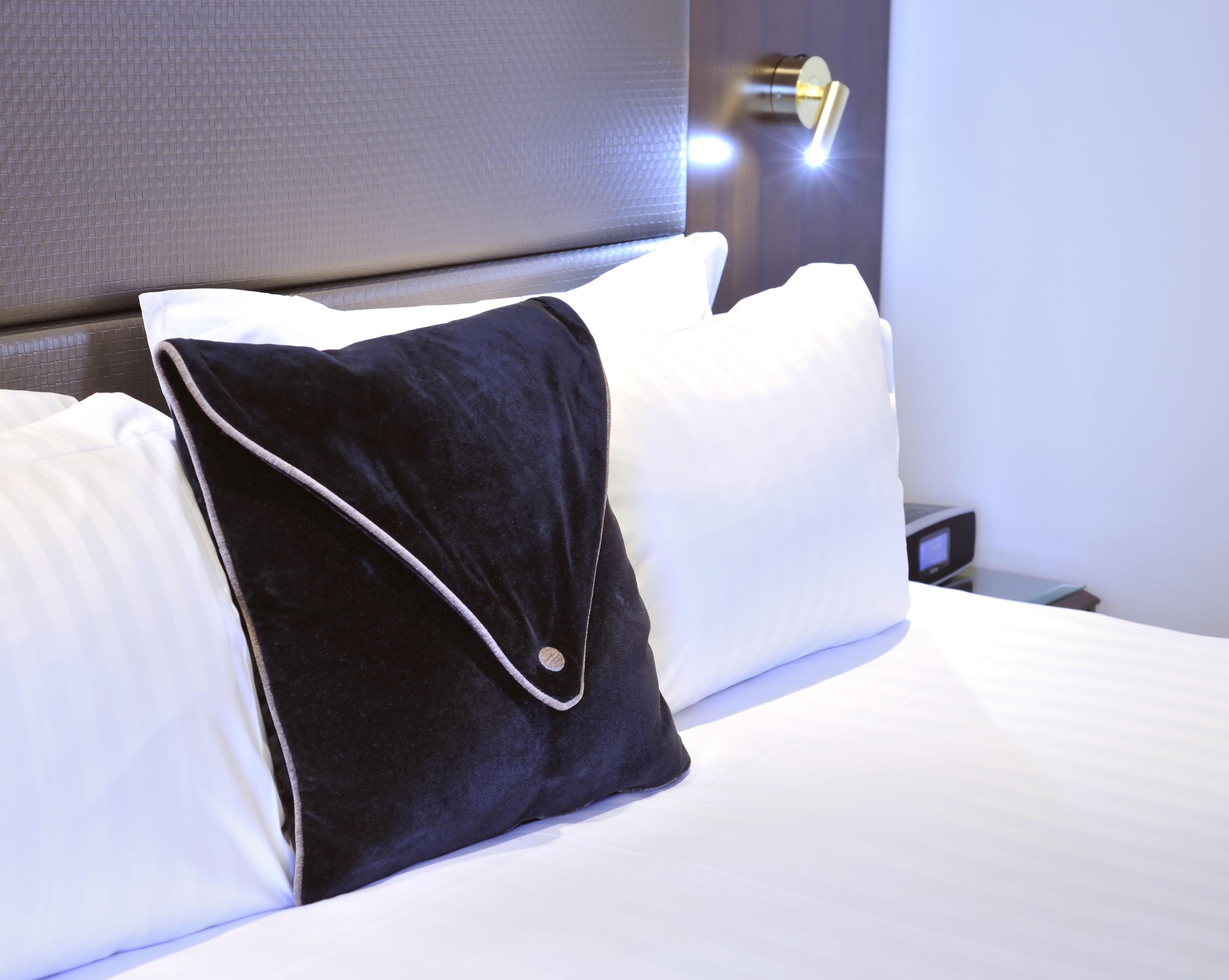מלון Stamford Plaza מלבורן מראה חיצוני תמונה The photo shows a neatly arranged bed with white linens. There are several pillows, with one black pillow featuring a distinctive design on top of the white pillows. The headboard of the bed is visible, along with a stylish wall-mounted light fixture
