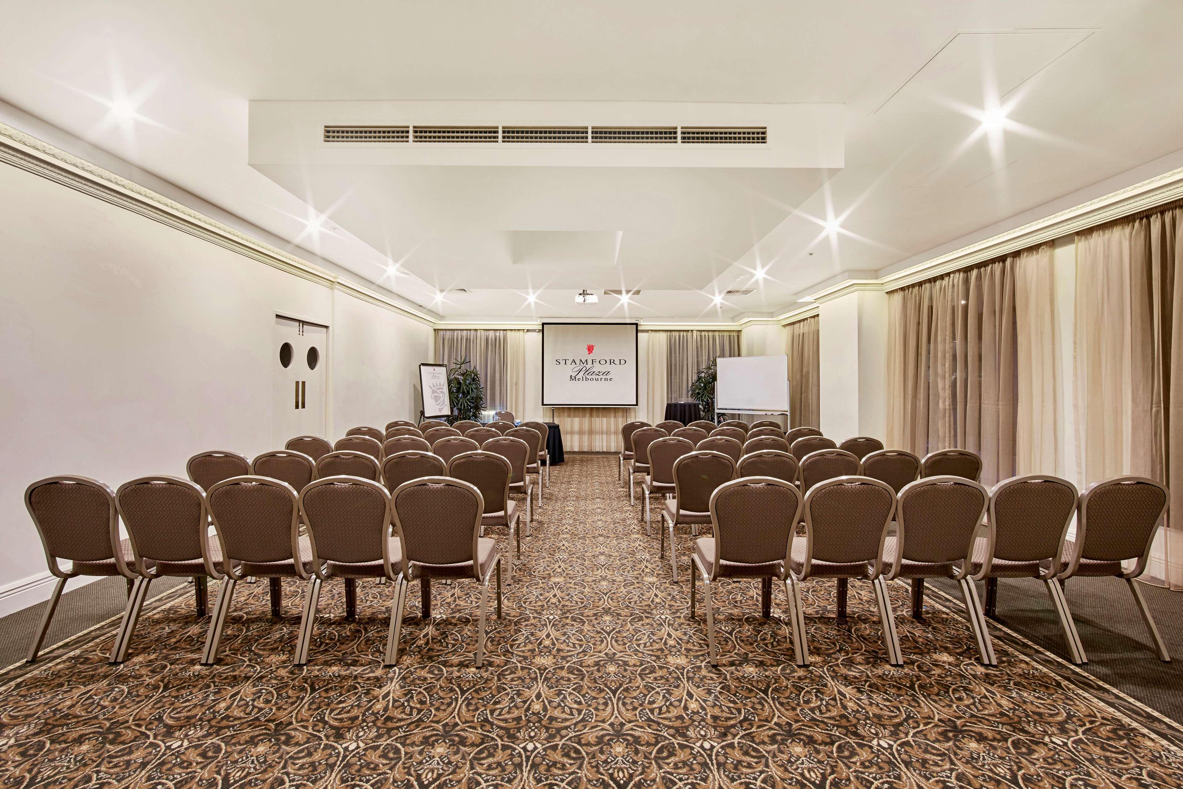מלון Stamford Plaza מלבורן מראה חיצוני תמונה The photo shows a conference room set up for an event. It features several rows of beige chairs arranged facing a central presentation area. At the front, there is a screen displaying a logo or title, possibly for a conference or meeting. The walls h