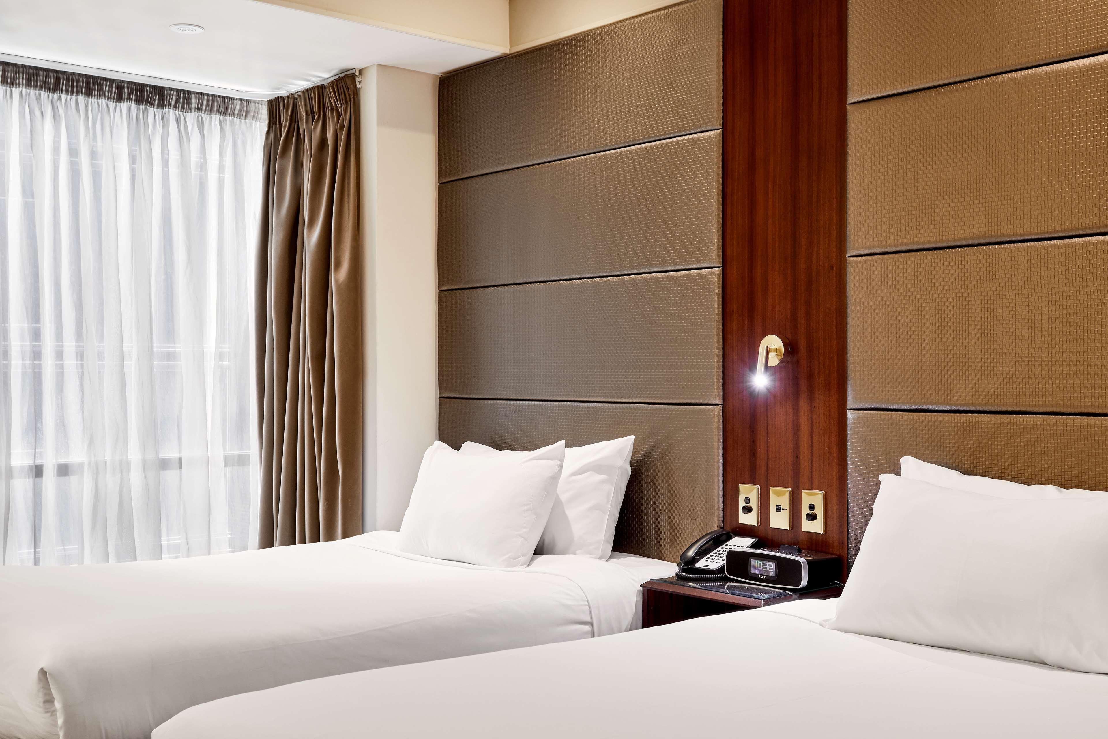 מלון Stamford Plaza מלבורן מראה חיצוני תמונה The photo shows a modern hotel room featuring two twin beds with white linens and fluffy pillows. The walls behind the beds have a combination of textured panels in neutral colors. There is a bedside table with a lamp and a phone. A large window with