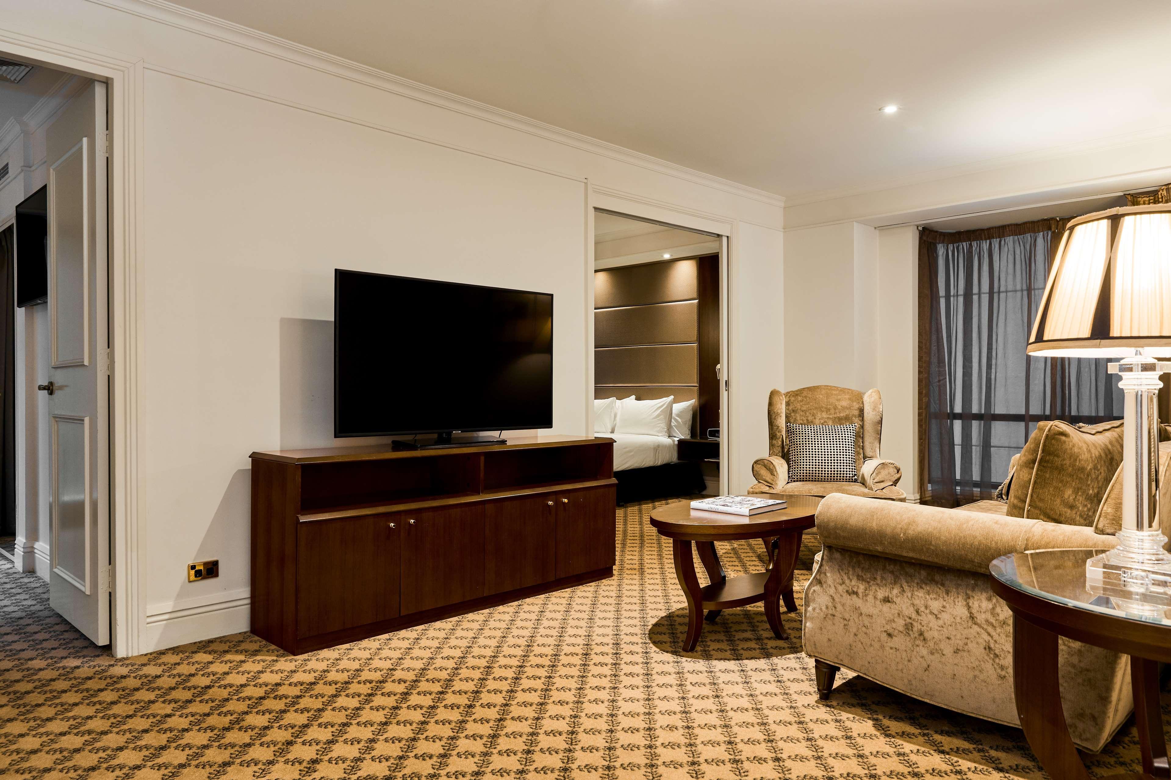 מלון Stamford Plaza מלבורן מראה חיצוני תמונה The photo shows a well-furnished room, likely a hotel suite or a living area. There is a large flat-screen television mounted on a wooden entertainment unit. In front of the TV, there is a round wooden coffee table. To the side, a comfortable armchai