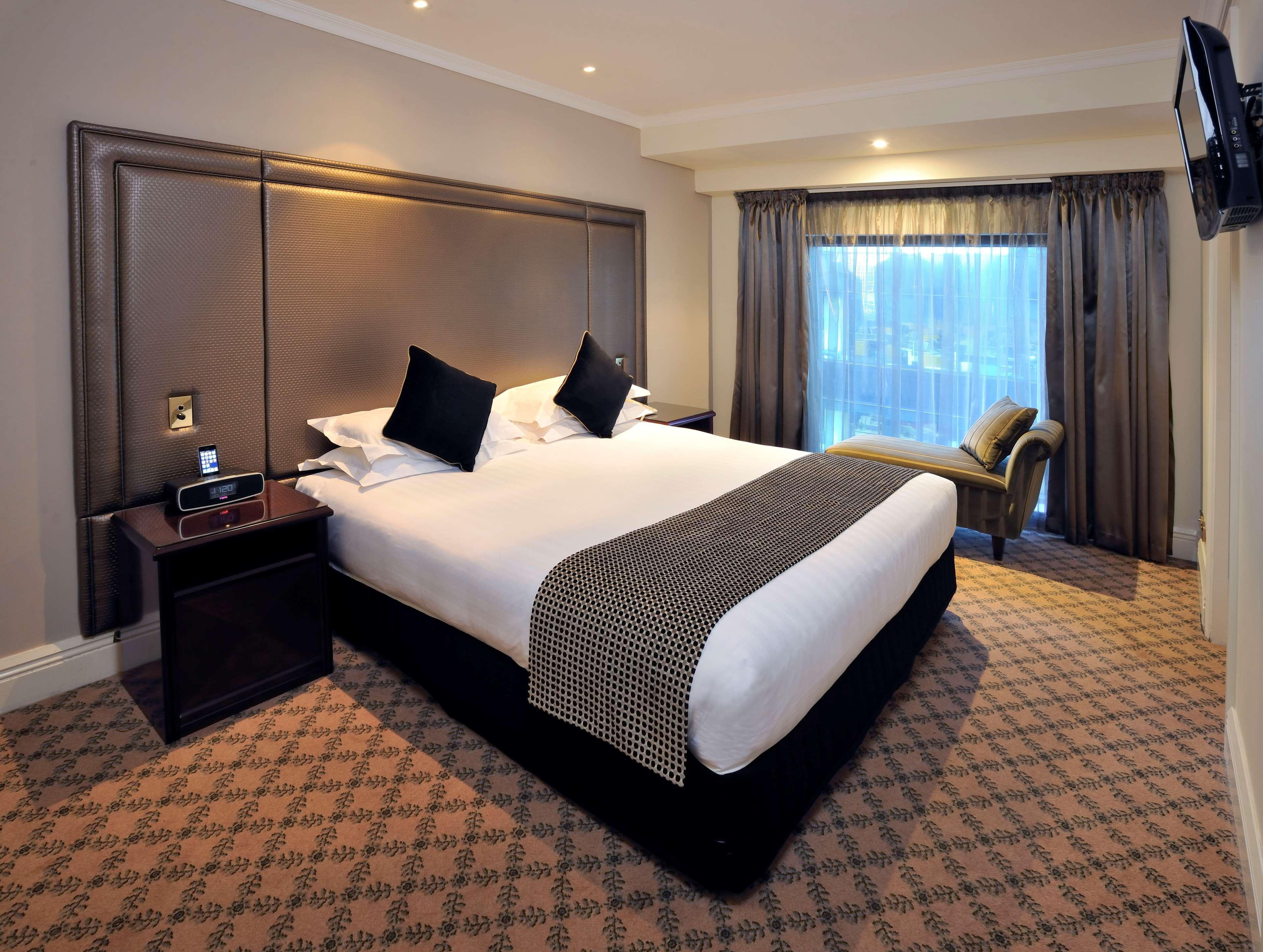 מלון Stamford Plaza מלבורן מראה חיצוני תמונה The photo shows a hotel room featuring a neatly made king-sized bed with dark pillows. The headboard is upholstered in a dark material, and there is a decorative blanket at the foot of the bed. Beside the bed are two nightstands, each equipped with l