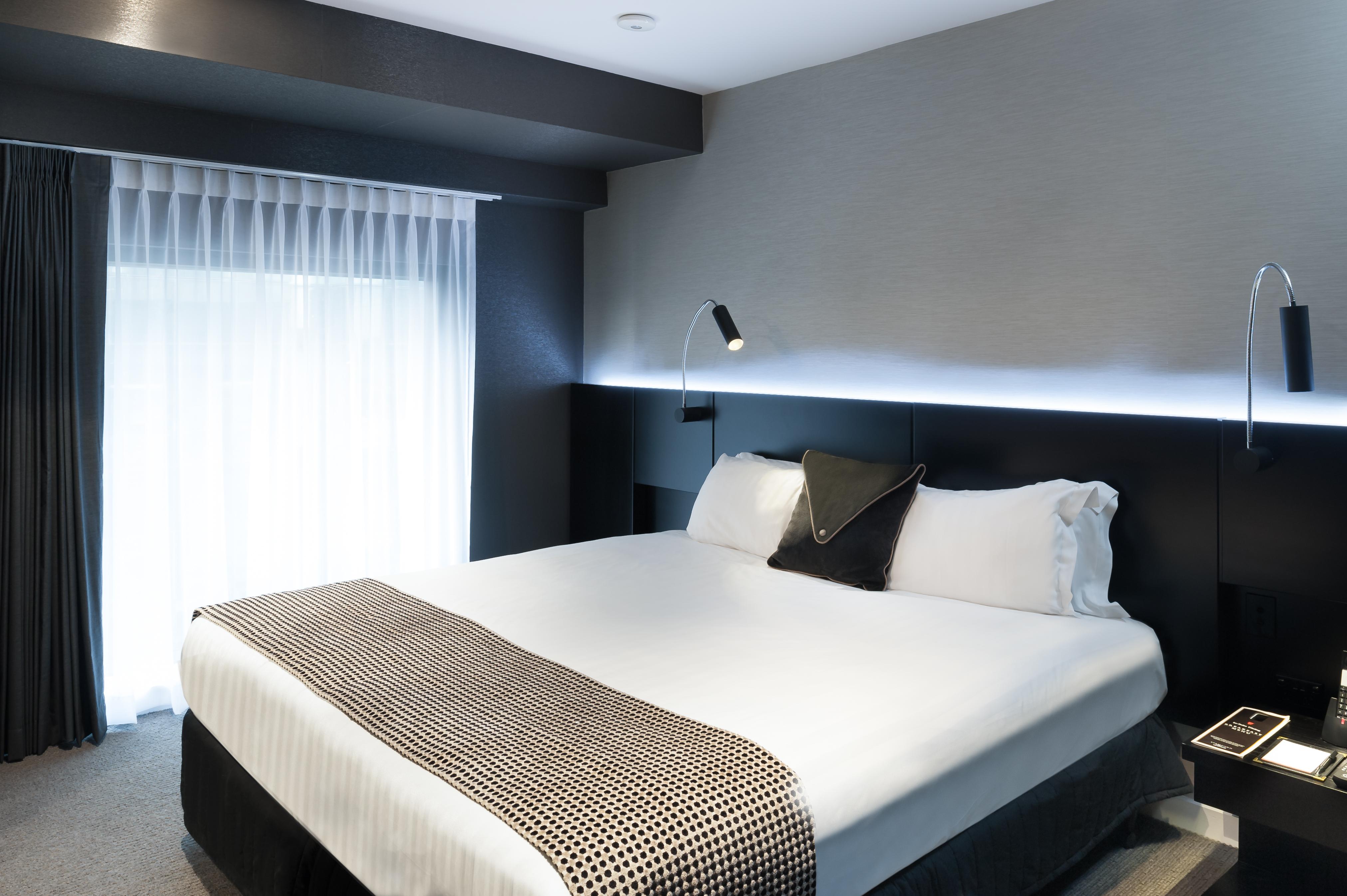 מלון Stamford Plaza מלבורן מראה חיצוני תמונה The photo shows a modern hotel room featuring a large bed with white linens and a decorative throw blanket. The room has a sleek and contemporary design, with gray walls and a soft lighting strip along the headboard. A window is adorned with sheer cu