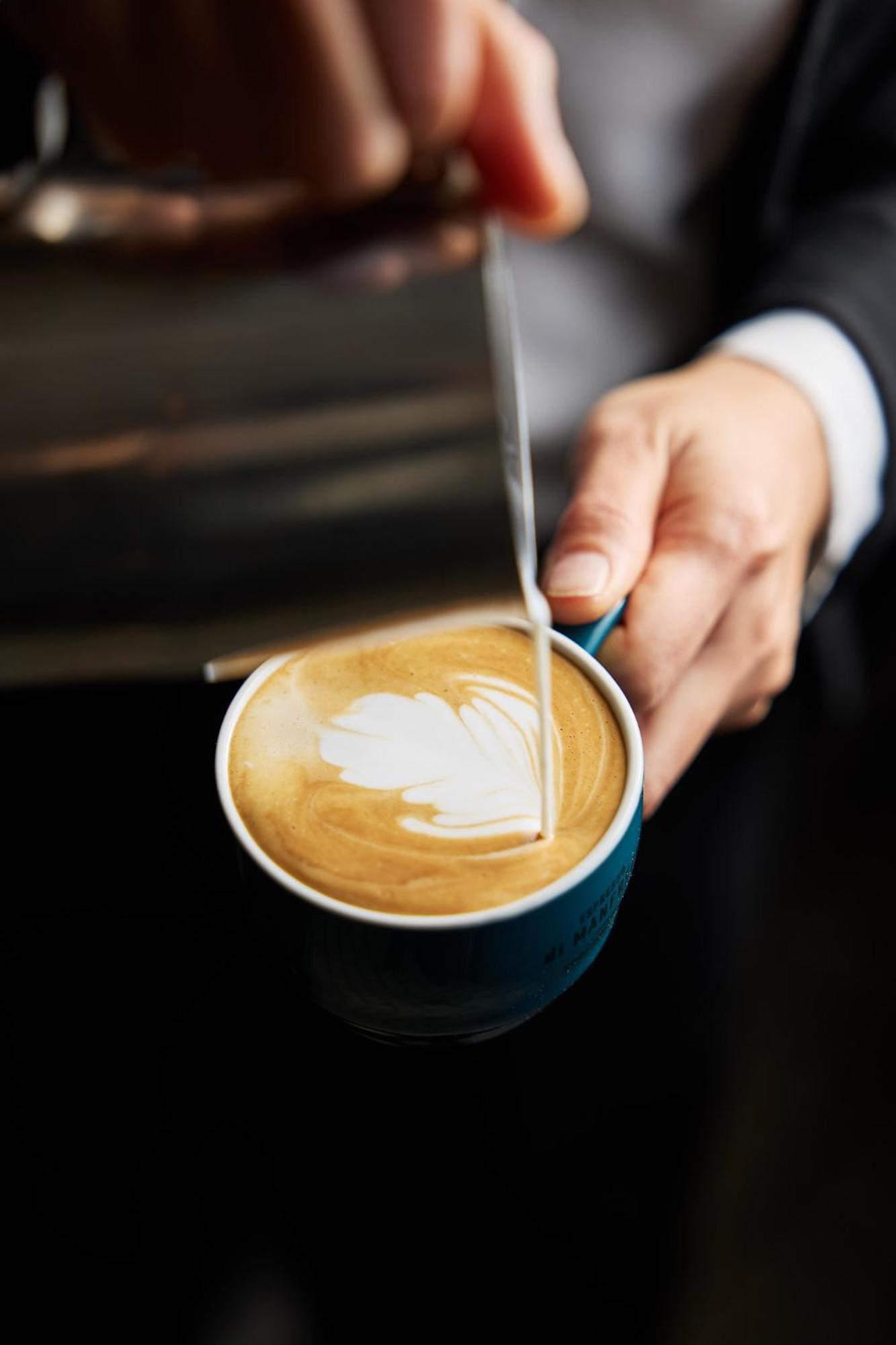 מלון Stamford Plaza מלבורן מראה חיצוני תמונה The photo shows a close-up of a hand holding a cup of coffee with a latte art design on the surface. The coffee appears to be a creamy, caramel color, likely a latte or cappuccino. A small stream of milk is being poured into the cup, creating a heart