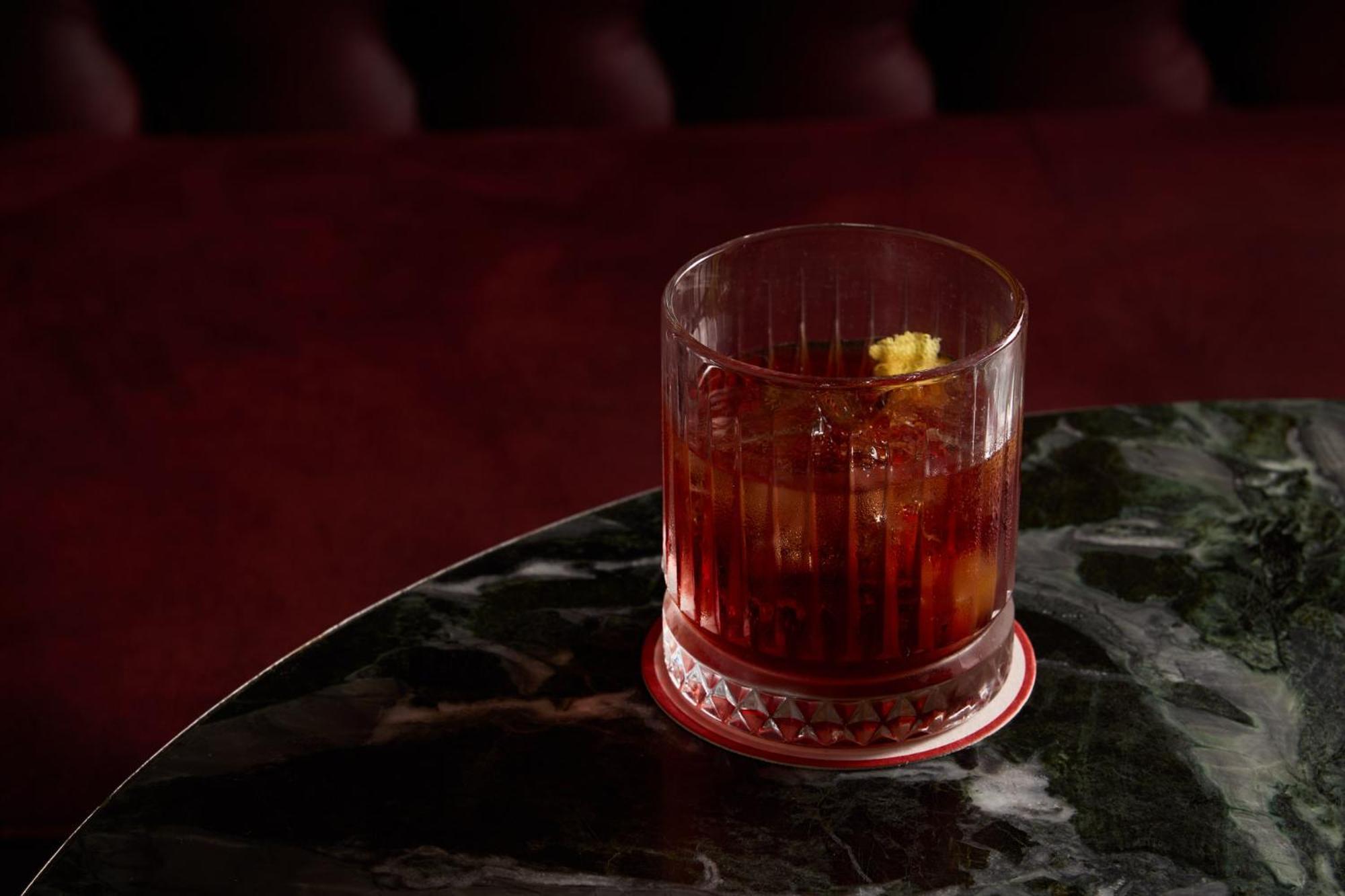 מלון Stamford Plaza מלבורן מראה חיצוני תמונה The photo depicts a short glass filled with a dark-colored beverage, likely an alcoholic drink. The glass has a textured design, with ice cubes visible inside. A twist of lemon peel is perched on the rim, adding a touch of garnish. The drink sits on 