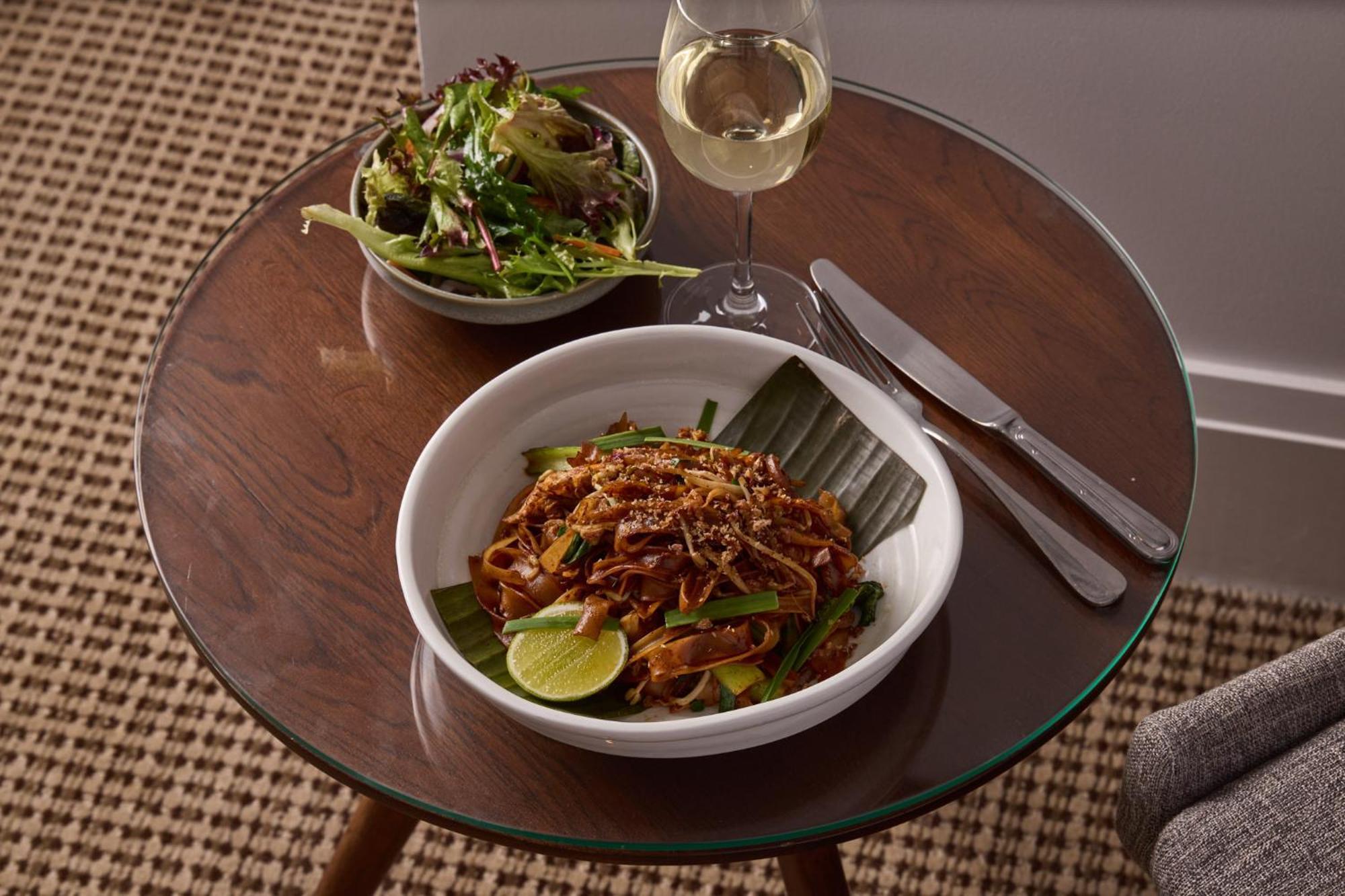מלון Stamford Plaza מלבורן מראה חיצוני תמונה The photo shows a meal served on a wooden table. In the foreground, there is a bowl of stir-fried noodles garnished with green onions and a lime wedge, set atop a banana leaf. Next to it, there is a bowl of mixed green salad. A glass of white wine si